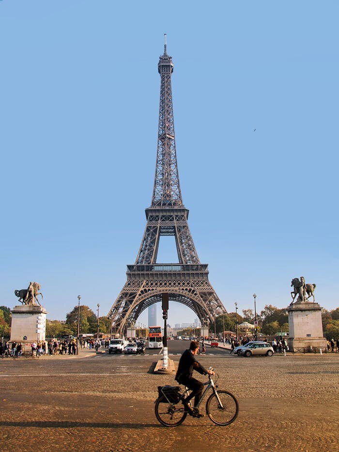 Bicyclist rides past the iconic Eiffel Tower in Paris under a clear blue sky.